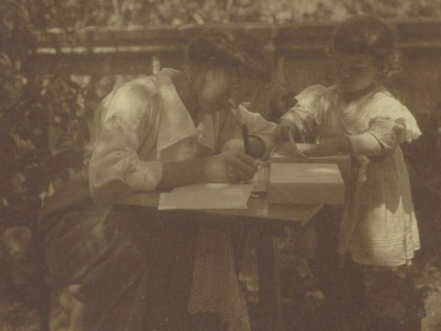 Fotografie welche Helene von Wild an einem niedrigen Tisch im Freien am Schreiben zeigt. Neben ihr steht ein Kleinkind, welches ein Papier vom auf dem Tisch liegenden Papierstapel hebt.