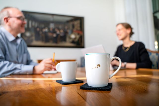 Zwei Kaffeetassen stehen auf einem Tisch. Im Hintergrund sind zwei Personen zu sehen, welche sich angeregt unterhalten.