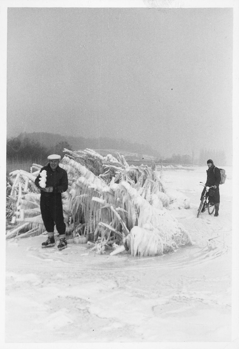 Fotografie zeigt ein zugefrorenes Gewässer auf welchem eine Person mit Schlittschuhen posiert und eine Person mit dem Fahrrad unterwegs ist.