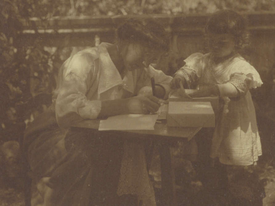 Fotografie welche Helene von Wild an einem niedrigen Tisch im Freien am Schreiben zeigt. Neben ihr steht ein Kleinkind, welches ein Papier vom auf dem Tisch liegenden Papierstapel hebt.